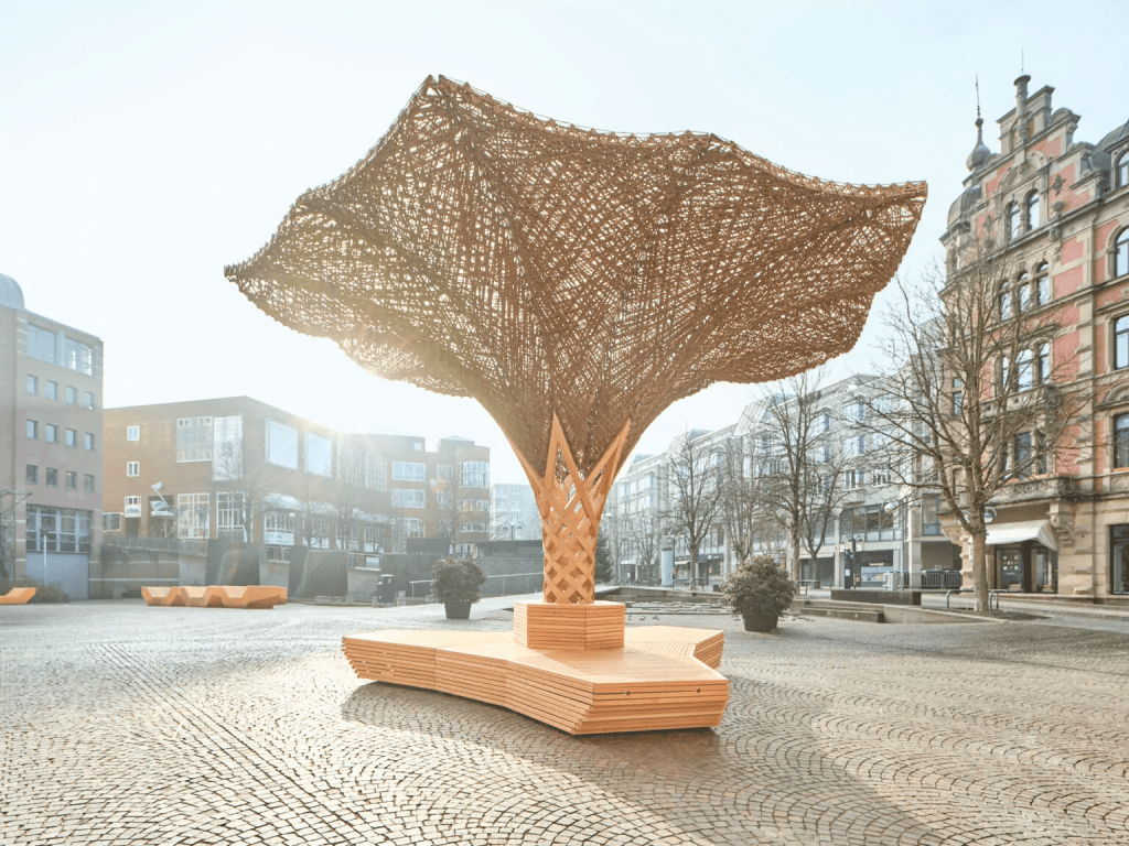 Skulptur aus Holz mit großem, kapselförmigen Netzdach in einem urbanen Platz, umliegende Gebäude.